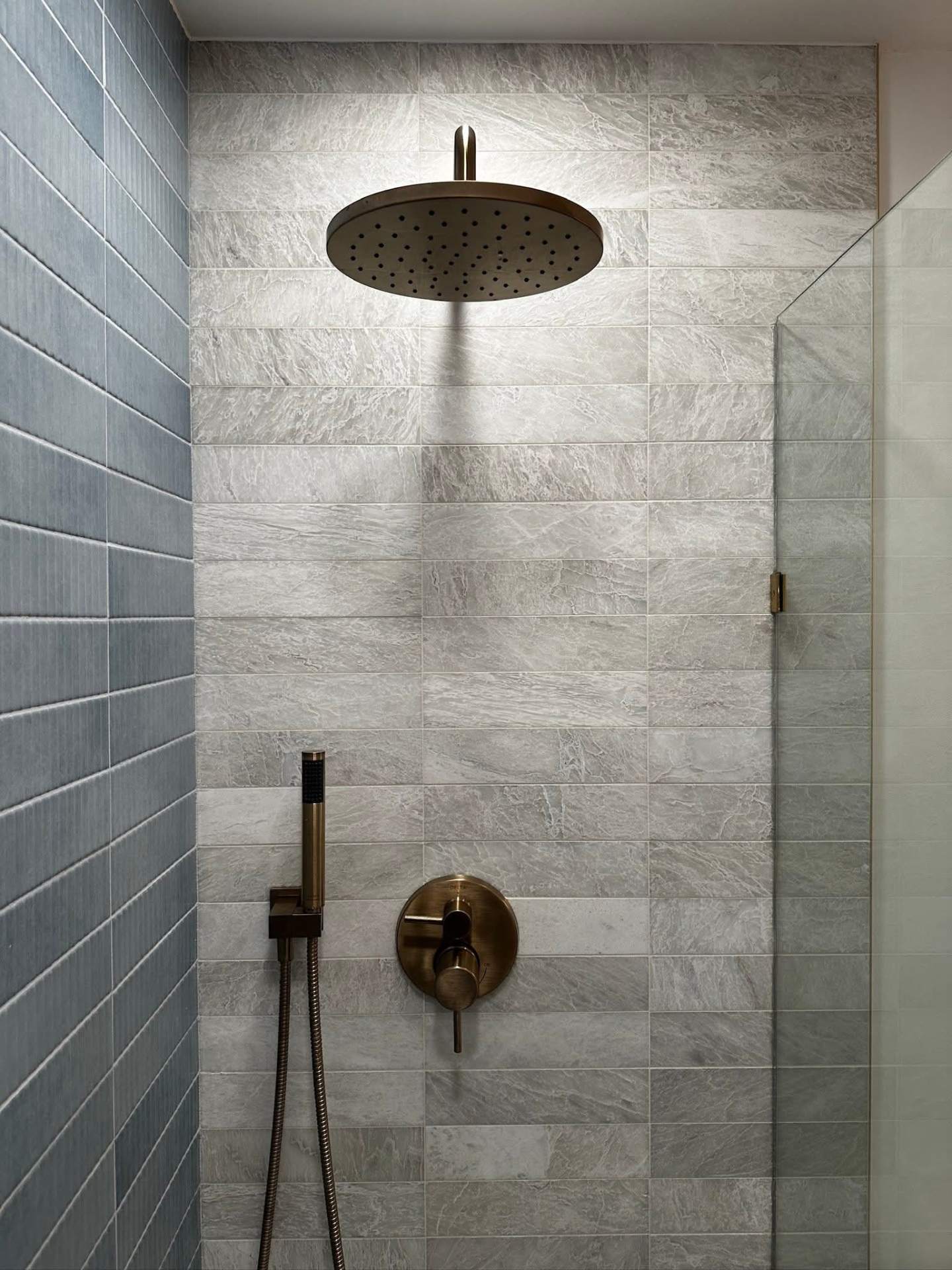 Brass rain shower head and hand shower on grey stone tile with blue accent wall, New York bathroom remodel