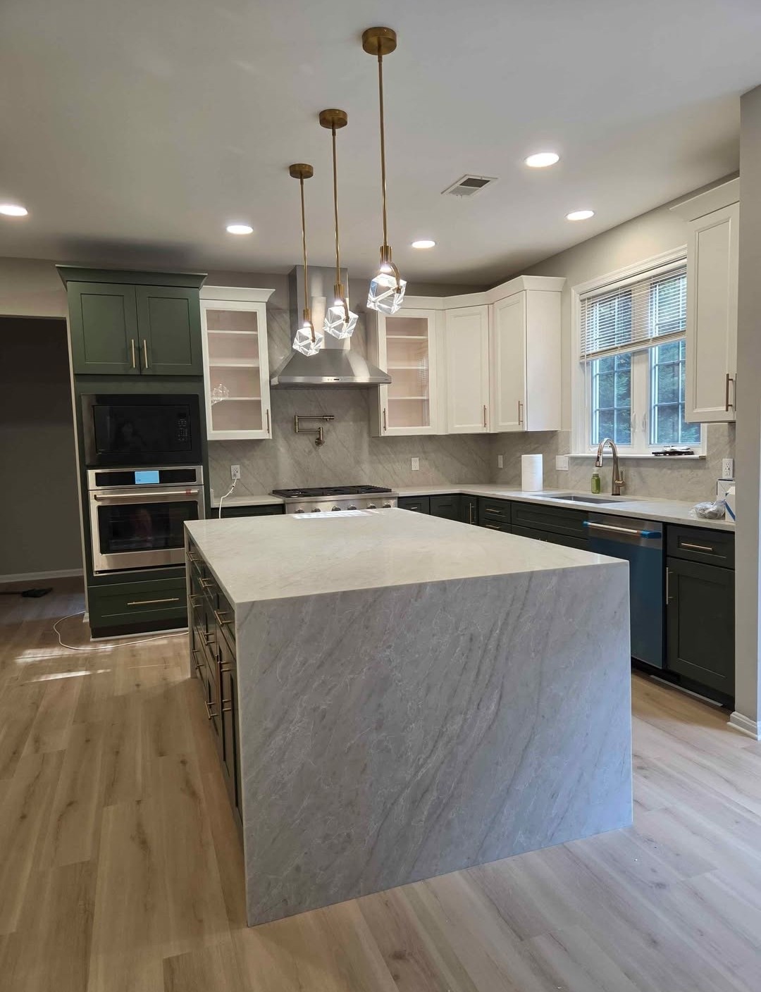 Renovated kitchen with waterfall marble island, green cabinetry and brass pendant lighting