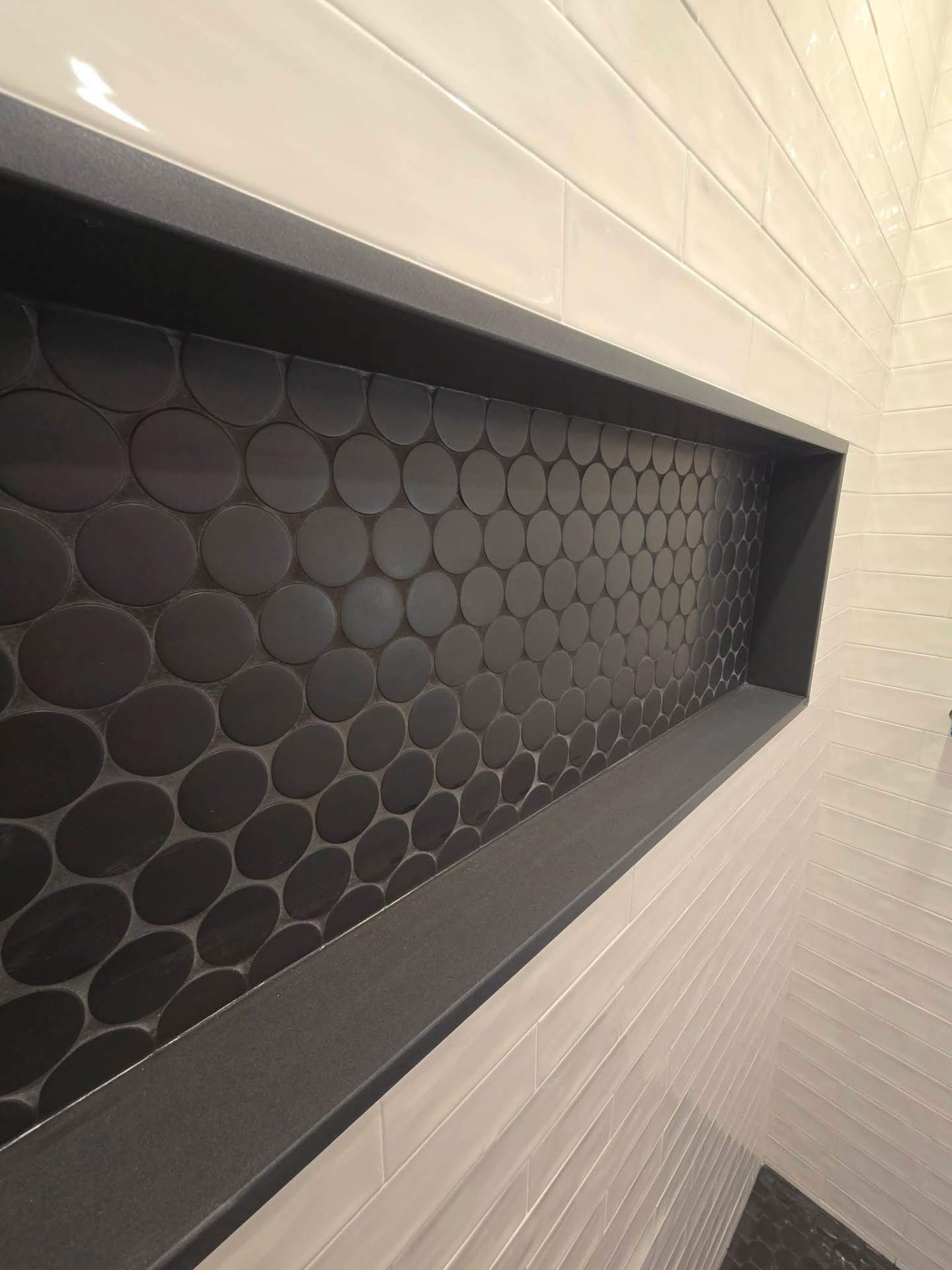 Close-up of a black penny-round tile shower niche with stone sill in a custom NYC bathroom